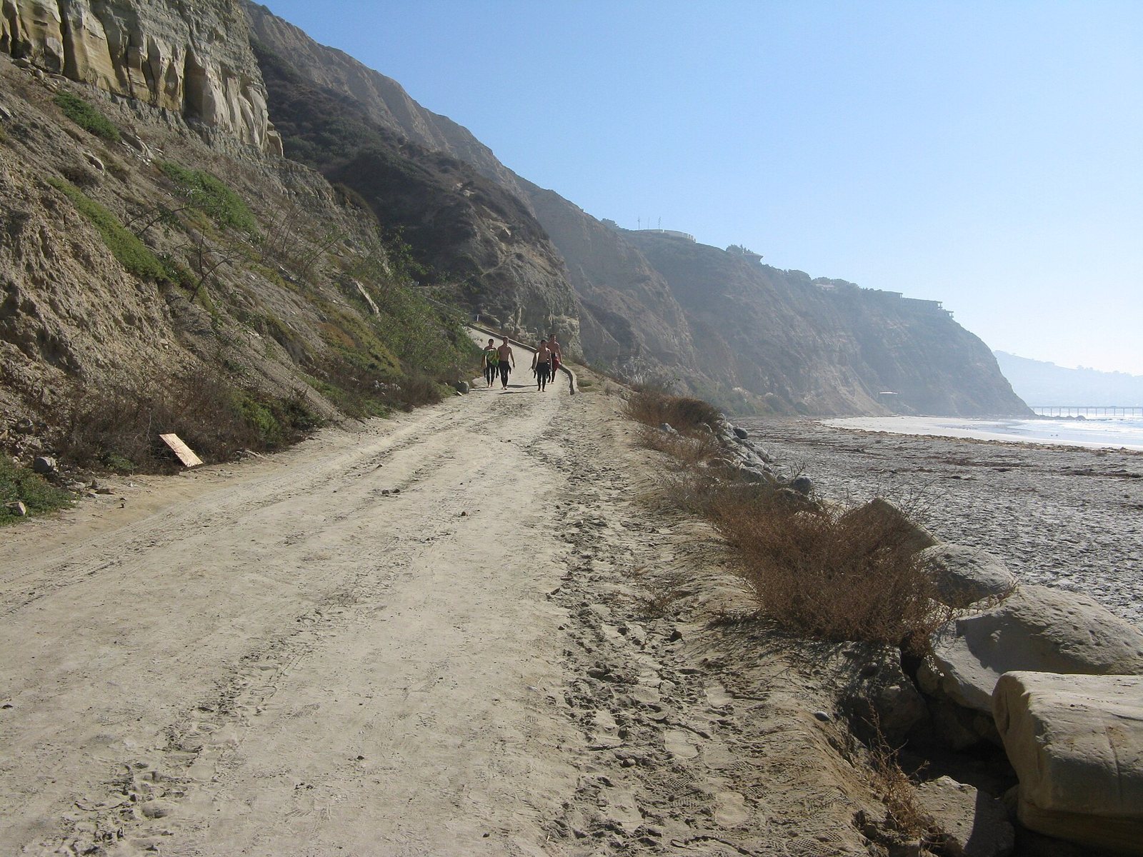 A dirt road running down beside steep sandstone cliffs toward the beach, a small group of people walking down it