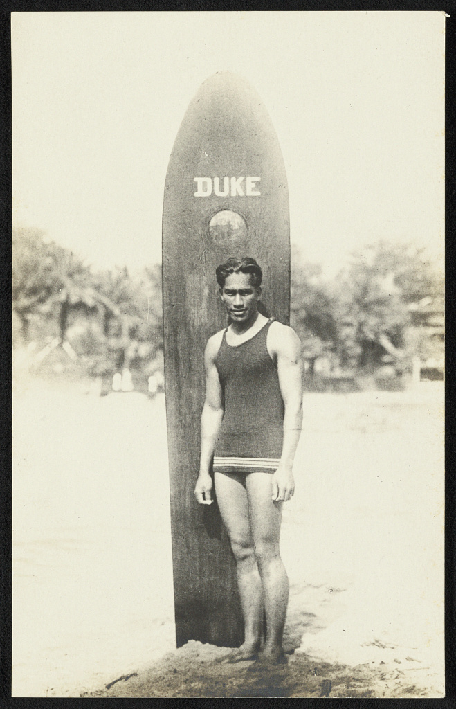 A young Duke Kahanamoku standing barefoot on sand in a dark one piece swimsuit, in front of a tall solid wooden surfboard stood upright behind him with the word DUKE painted near its top.