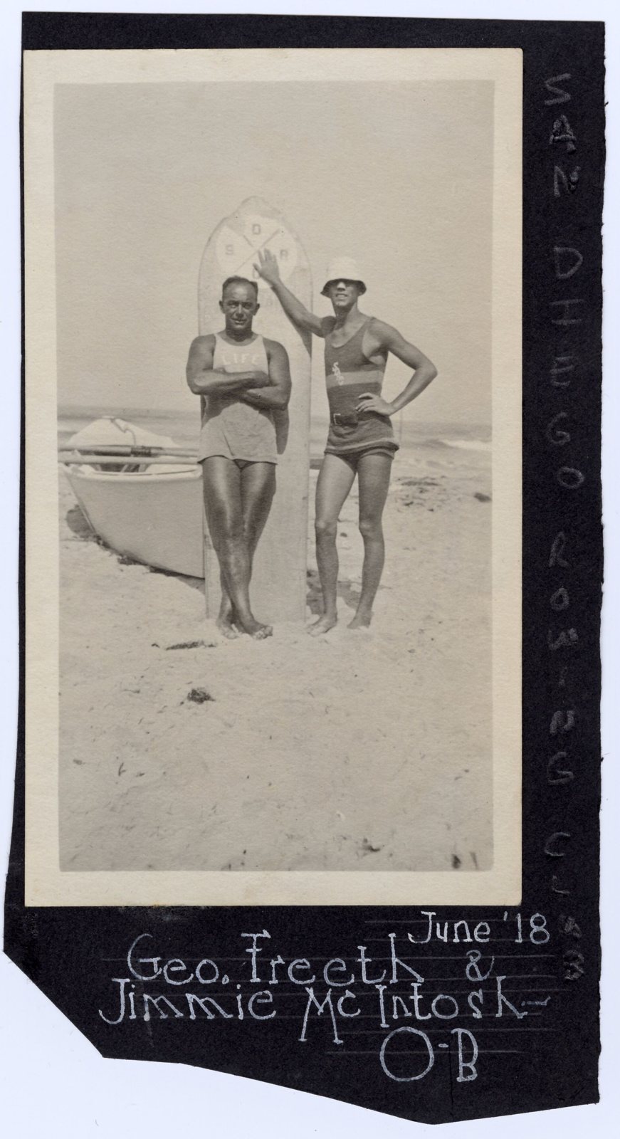 Two men in one piece wool swimsuits standing on the sand in front of a tall surfboard, arms crossed, a rowboat behind them, mounted on a dark album page lettered in white ink
