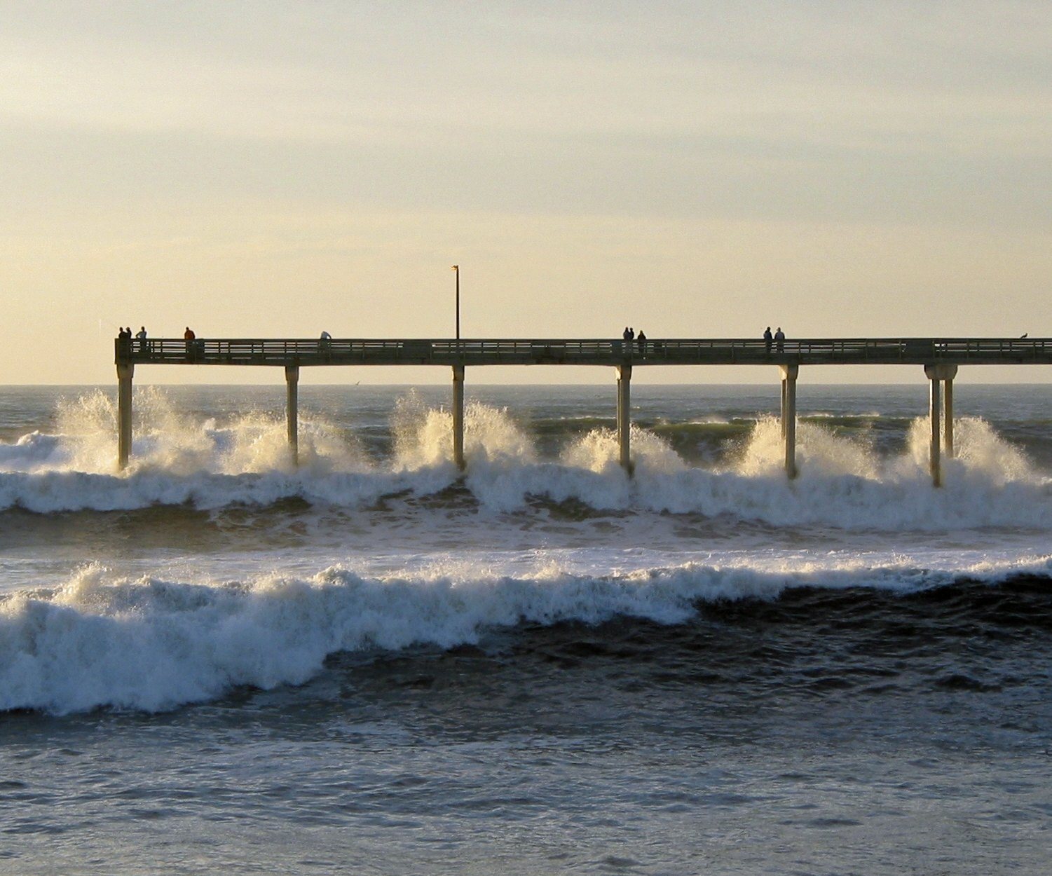 Waves breaking white under the long concrete Ocean Beach pier, people standing along its rail