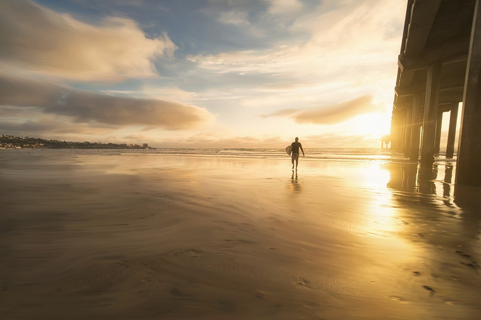 Silhouette of a surfer carrying a board across wet sand at sunset, the low sun breaking through clouds beside the pilings of a pier on the right