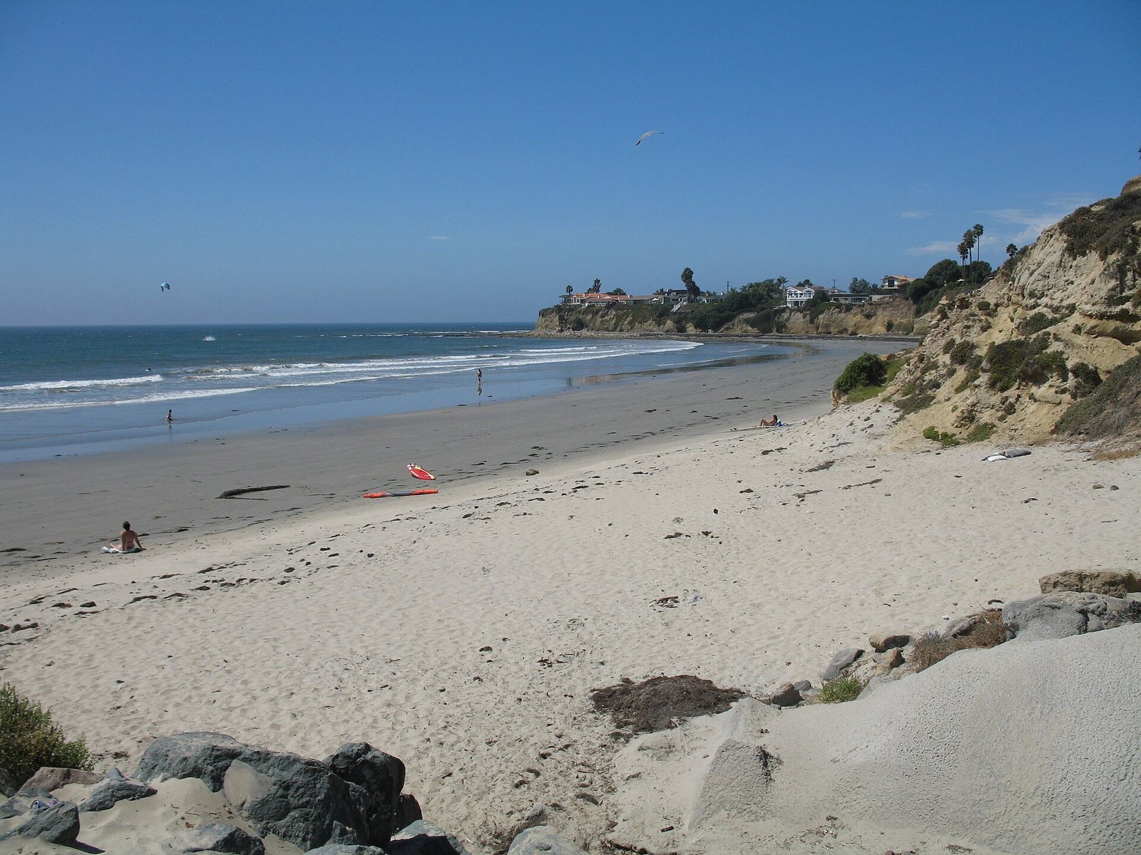 A wide sandy cove below low bluffs and a canyon mouth, a few people near the water under a clear sky