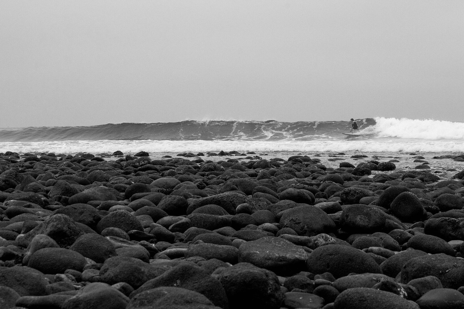 A beach of rounded cobblestones in the foreground and a single surfer riding a long wave in the distance, in black and white