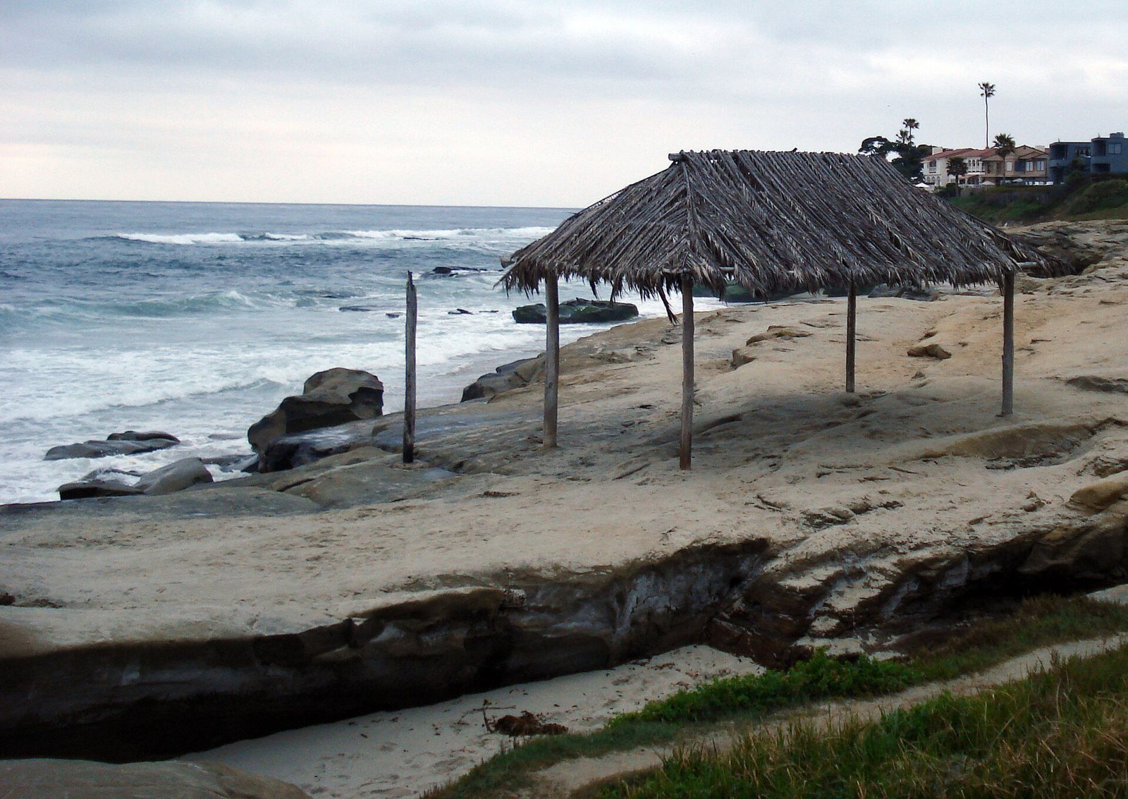 A palm thatched shade shelter on wooden poles on a sandstone ledge above the surf, houses on the bluff behind