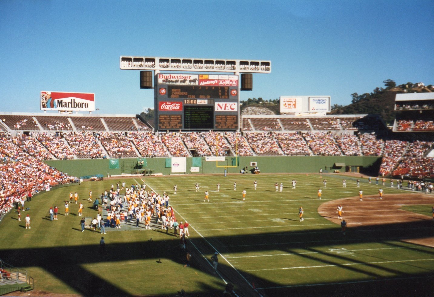 A football game seen from the upper stands of a packed stadium, players and a crowded sideline on the grass, a tall scoreboard and billboards above the far seats under a clear blue sky