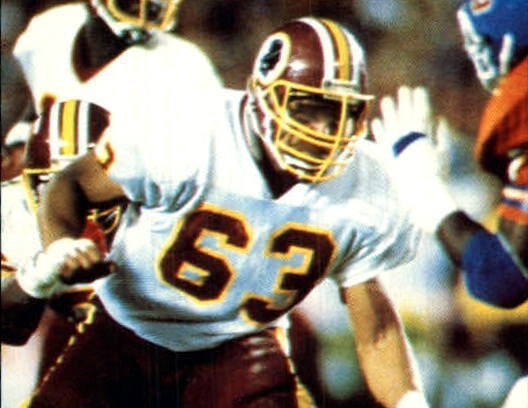 A football lineman in a white Washington jersey with burgundy number 63 and a burgundy helmet blocking in the middle of a play