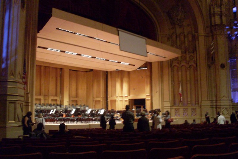 The inside of an ornate concert hall seen from the main floor, an orchestra stage under a wooden acoustic canopy with gilded walls on either side