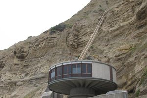 A round concrete house with a ring of windows sits on a pedestal at the foot of a steep sandstone cliff, behind a curved concrete wall above a rocky beach, with a narrow tram track climbing the cliff to a small landing at the top