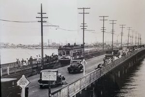 A wide wooden bridge over the water lined with utility poles, a streetcar and old automobiles crossing, people standing along the railing, and a road sign in the foreground