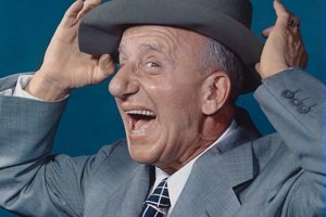 Color portrait of Jimmy Durante in a grey suit, tipping his hat back with both hands and laughing, photographed in 1948
