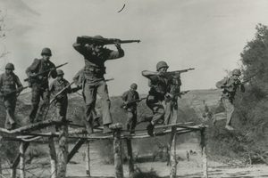 Marines in helmets carrying rifles run and leap along a raised course of narrow log beams over bare ground, brush and a hillside behind them
