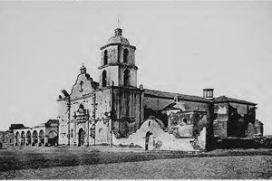 Mission San Luis Rey photographed in 1887, the bell tower and church front standing over walls that are visibly crumbling, with roof missing in places and the long cloister arcade running off to the left