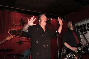A man with both hands raised and mouth wide open, singing onstage in front of a deep red curtain, with band members behind him