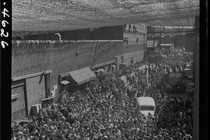 A street packed with thousands of workers walking toward the camera beneath a ceiling of patterned camouflage netting, a bus parked in the crowd
