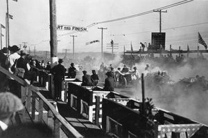 Racing cars pulling away in a cloud of dust under a Start and Finish banner, spectators behind a wooden rail on the left and a lap scoreboard on a pole at right