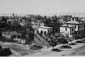 San Diego photographed in 1887 looking west toward the harbor, a windmill and water tower over scattered frame houses, young trees and a picket fence, with open dirt lots between them and Point Loma on the horizon