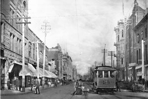 A wide downtown street with streetcar tracks down the middle, a trolley stopped at right, ornate brick and stone buildings with awnings on both sides and telegraph poles along the curb