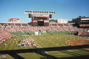 A football game seen from the upper stands of a packed stadium, players and a crowded sideline on the grass, a tall scoreboard and billboards above the far seats under a clear blue sky