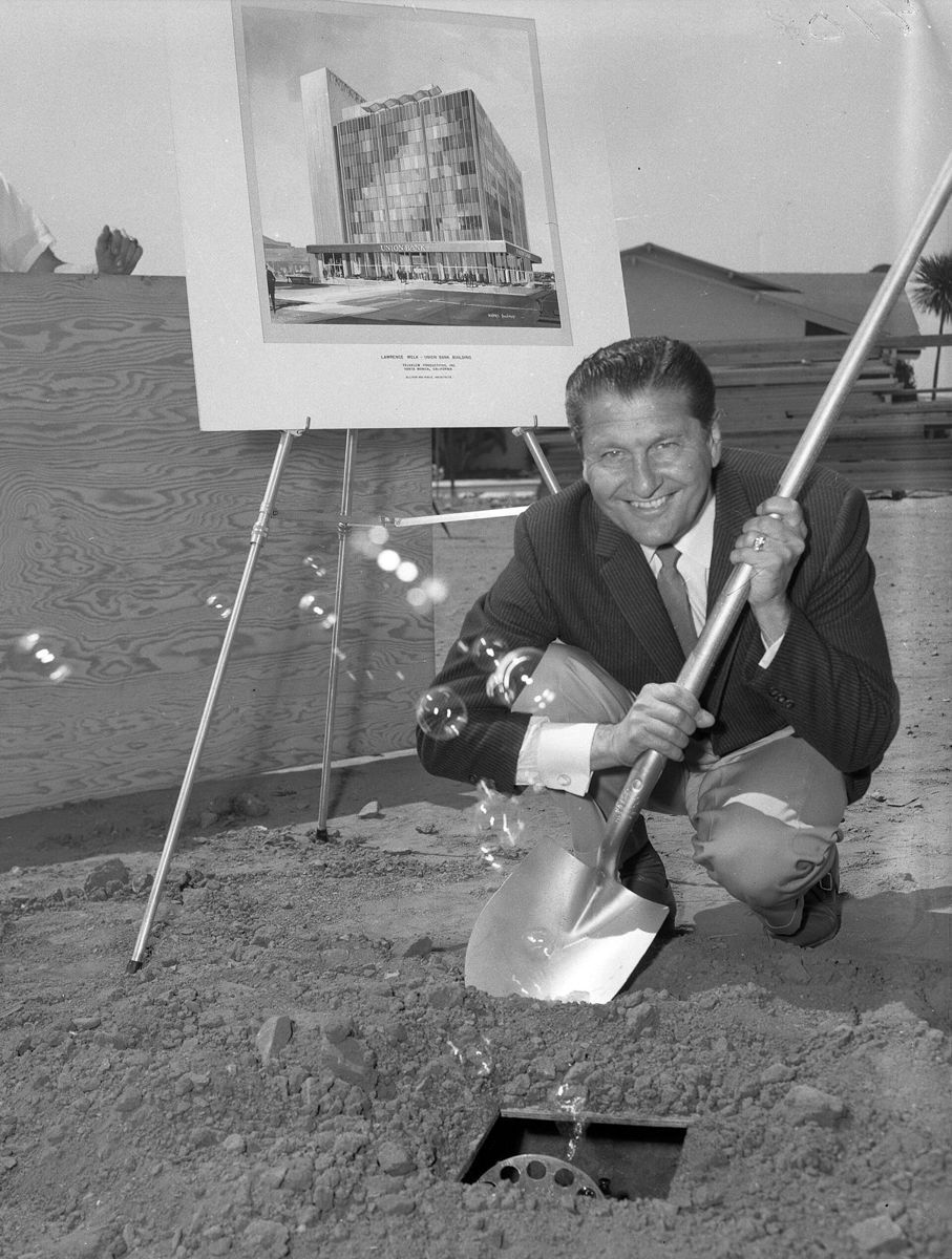 Lawrence Welk crouching and grinning with a shovel beside a small hole as bubbles float up, an architect's drawing of a building on an easel behind him