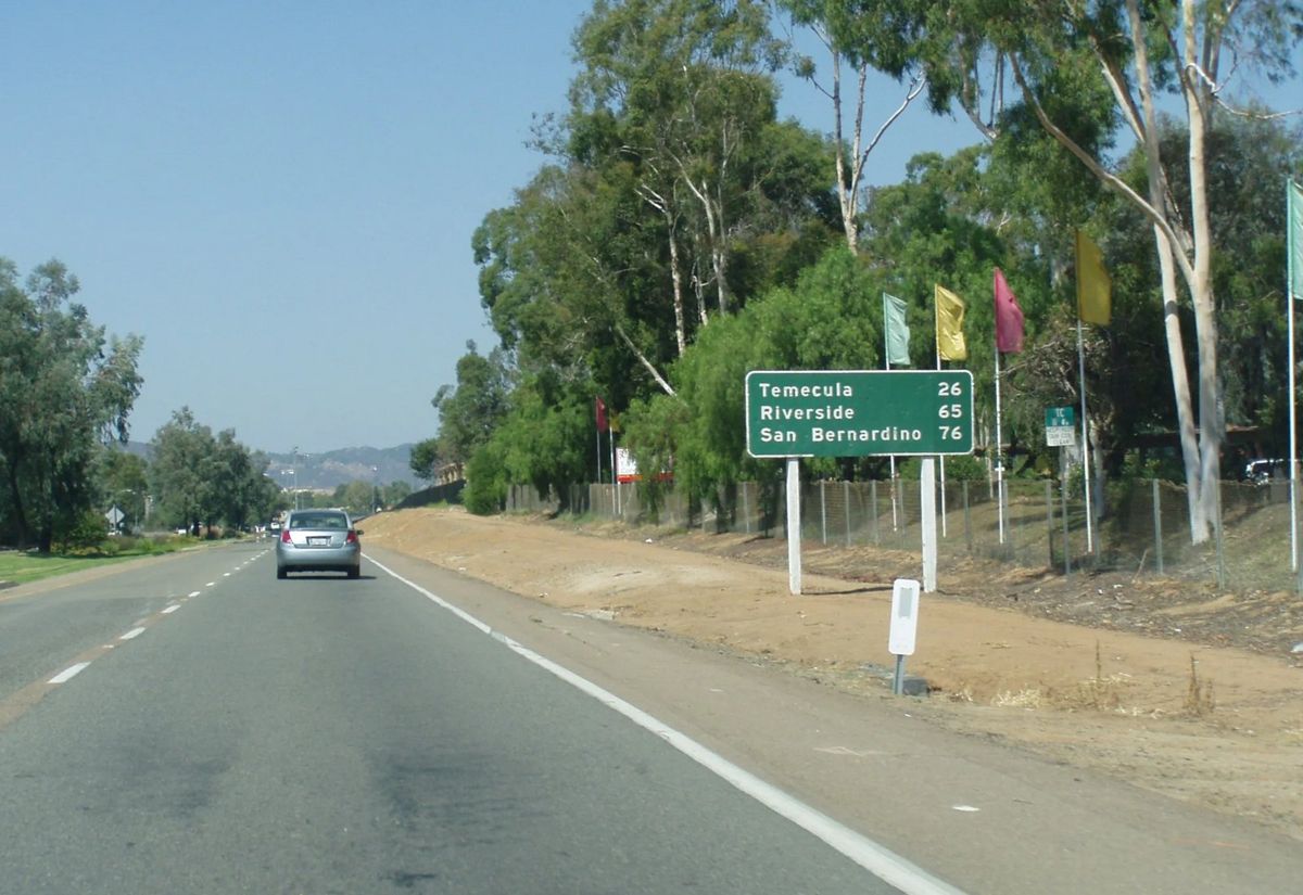 A two lane highway running north between eucalyptus trees, a green sign for Temecula, Riverside and San Bernardino at the right