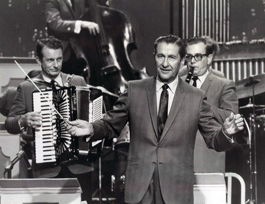 Lawrence Welk conducting in front of his orchestra on a television stage, an accordion player at left